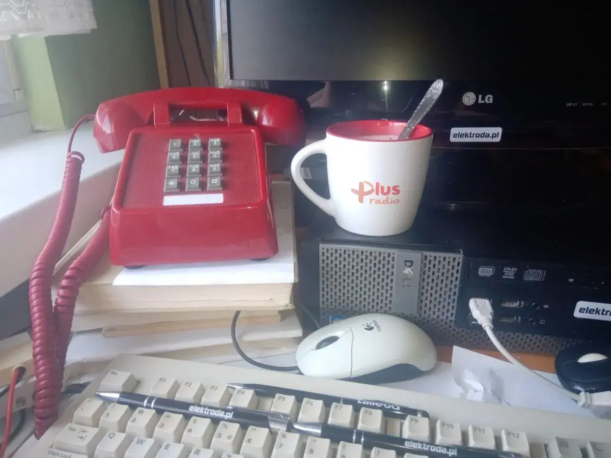 Czerwony telefon, kubek z napisem "Plus radio" i komputer na biurku.